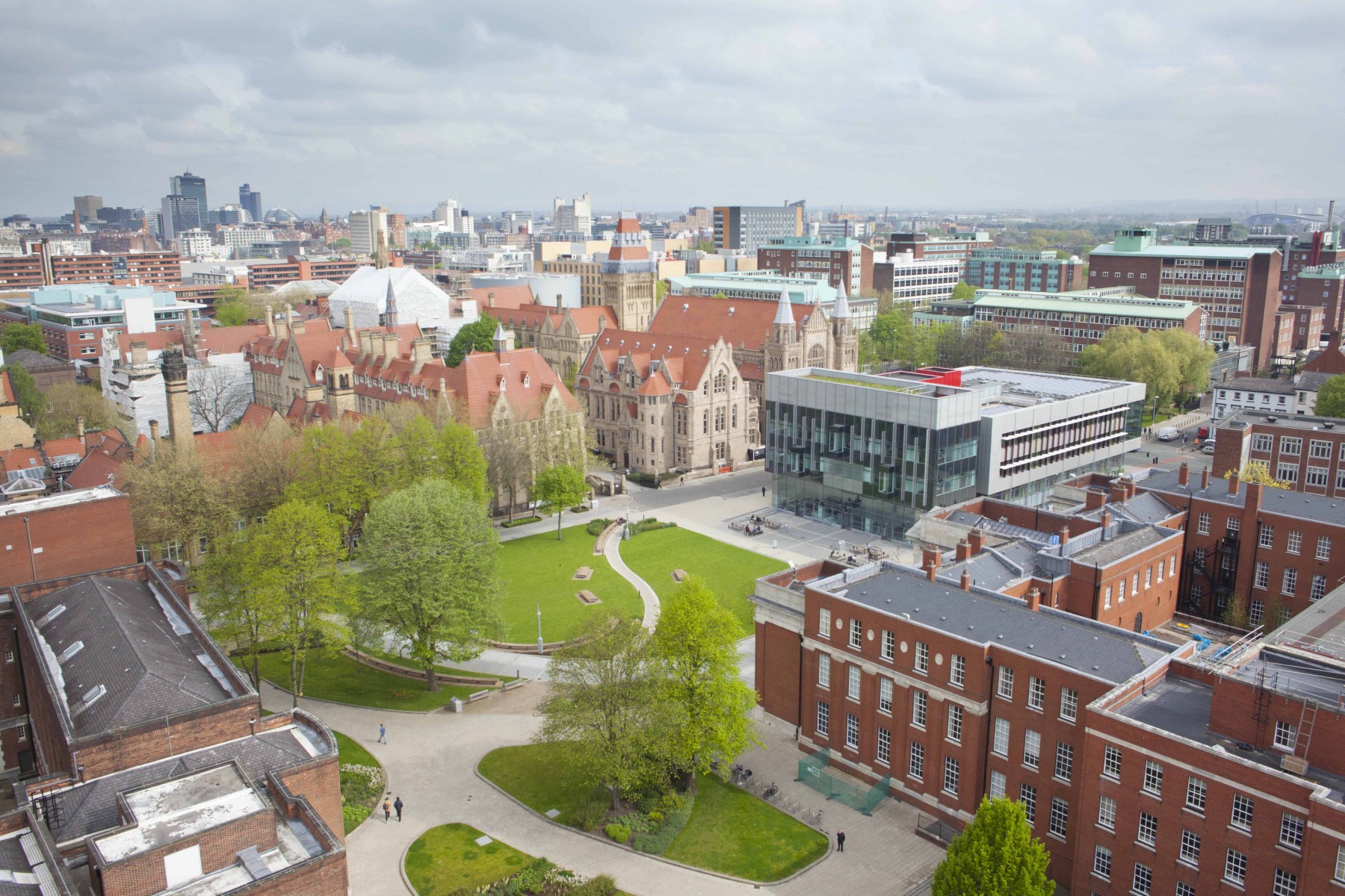 Aerial view of the University campus, showing Gilbert square, the modern architecture of the Learning Commons beside grand old buildings such as Whitwoth Hall and the tower. Manchester skyline in the background.